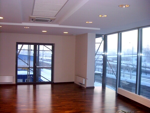 Wnętrze biura z dużymi oknami, parkietem w odcieniach brązu i białymi ścianami, widoczne grzejniki i elementy konstrukcyjne za oknami.